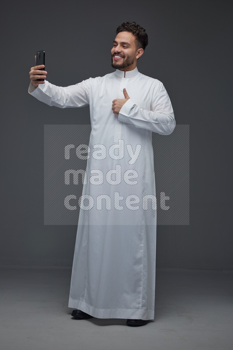A Saudi man wearing Thobe and taking selfie with his phone making different poses eye level on a gray background