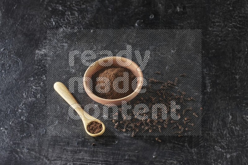 A wooden bowl and wooden spoon full of cloves powder with spreaded cloves on textured black flooring