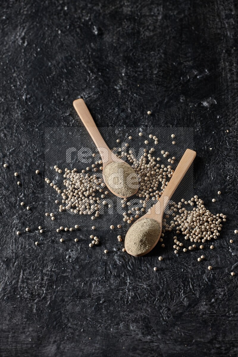2 wooden spoons full of white pepper powder with white pepper beads on textured black flooring
