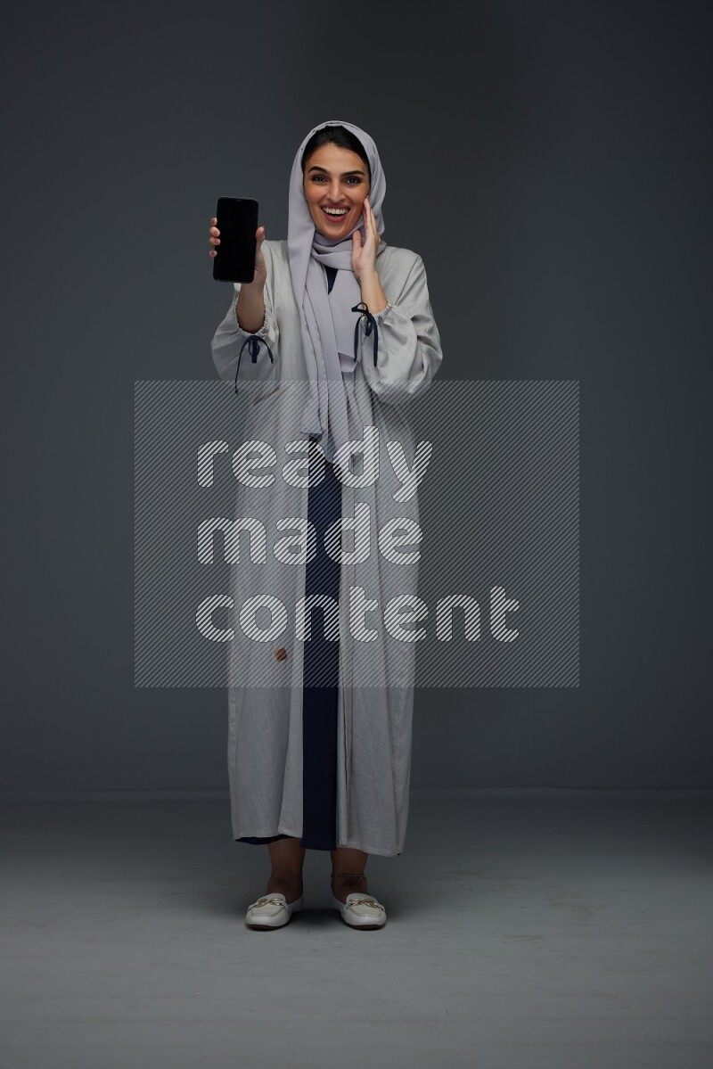 A Saudi woman wearing a light gray Abaya and head scarf standing and showing the phone's screen while holding a blue luggage eye level on a grey background