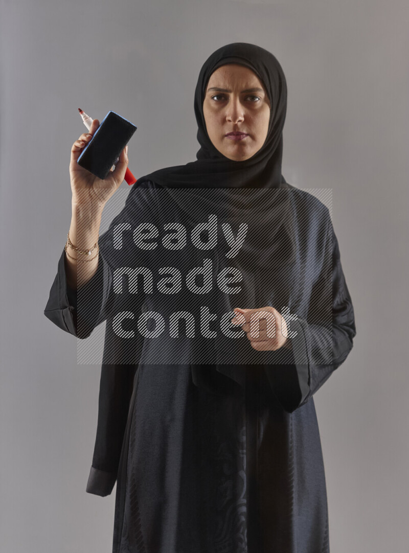 Woman in black abaya and hijab holding a marker pen to write on a transparent board with different reactions and expressions on grey background