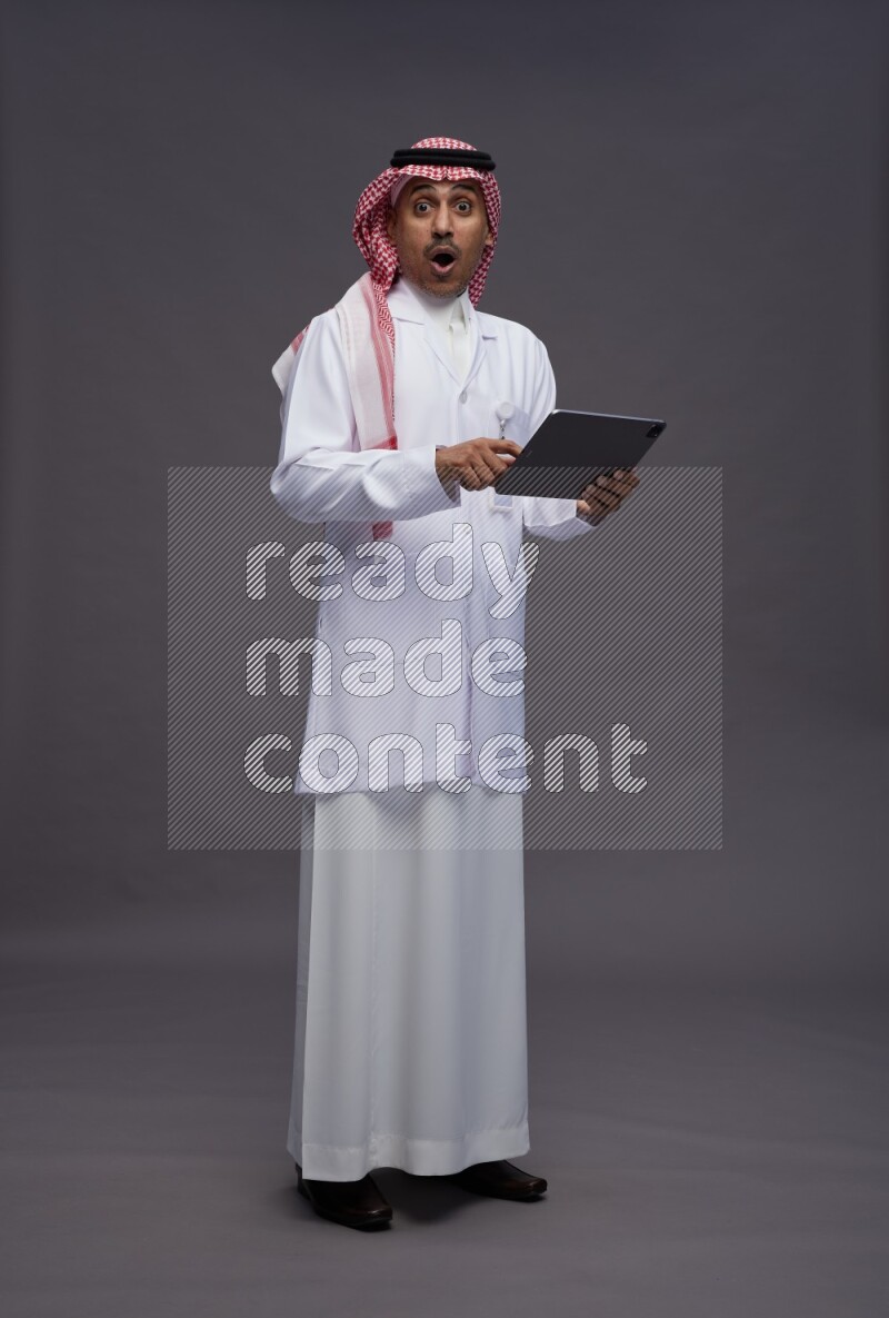 Saudi man wearing thob with lab coat and shomag with pocket employee badge standing working on tablet on gray background