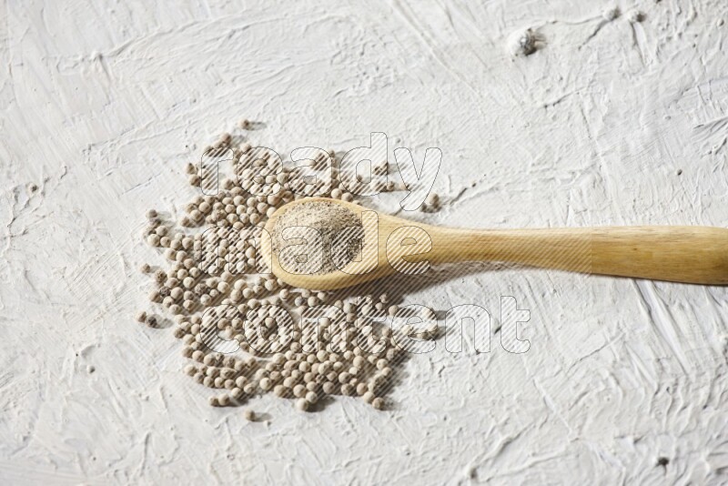 A wooden spoon full of white pepper powder and white pepper beads on textured white flooring