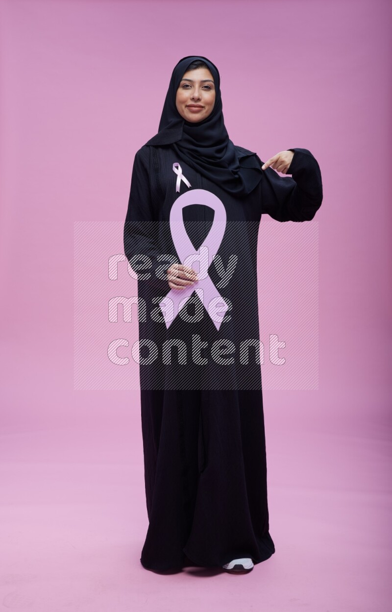 Saudi woman wearing pink ribbon on abaya standing holding awareness ribbon on plain pink background