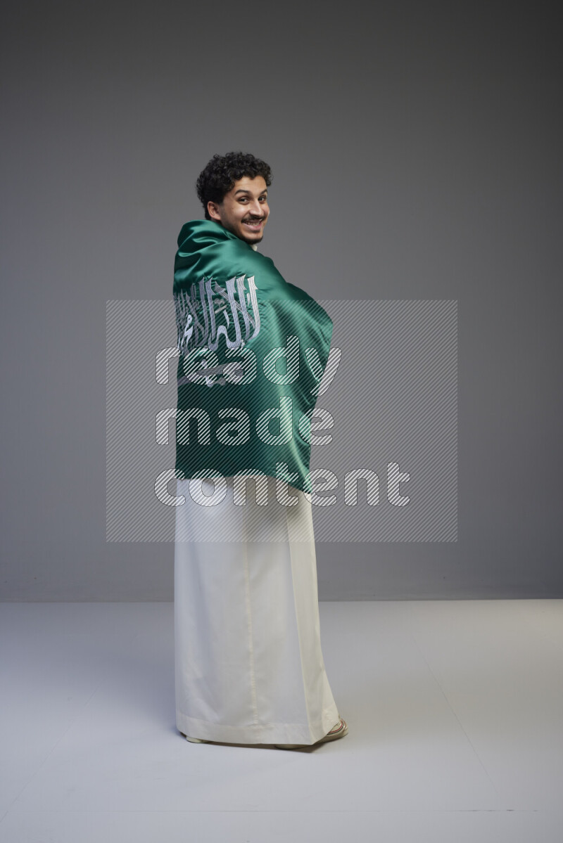 A Saudi man standing wearing thob wrapping big Saudi flag on gray background