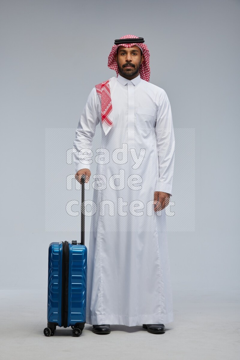 Saudi man wearing Thob and shomag standing holding Travel bag on Gray background