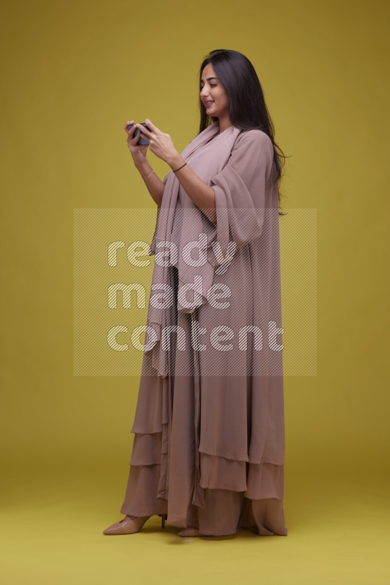 A woman Playing a Game on her smartphone on a Yellow Background wearing Brown Abaya