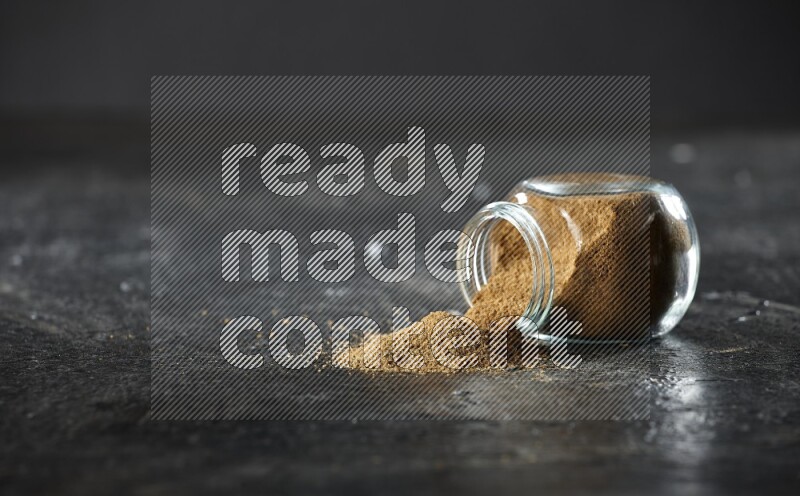 A flipped glass spice jar full of cumin powder and powder spilled out on a textured black flooring