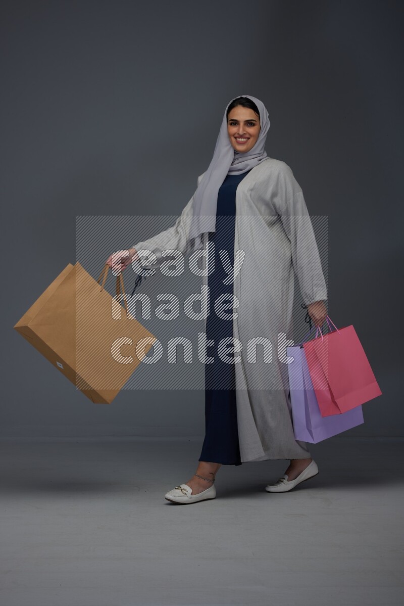 A Saudi woman wearing a light gray Abaya and head scarf standing and holding shopping bags on a grey background