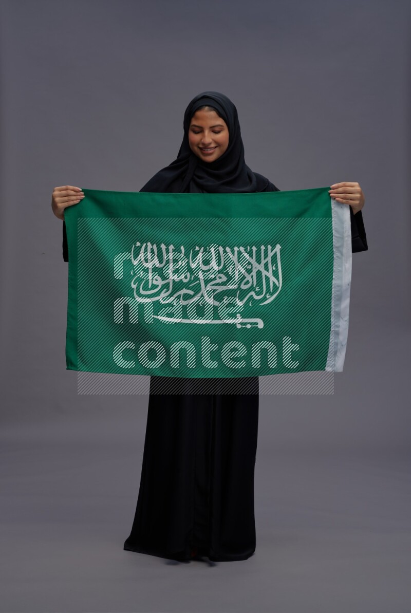 Saudi woman wearing Abaya standing holding Saudi flag on gray background