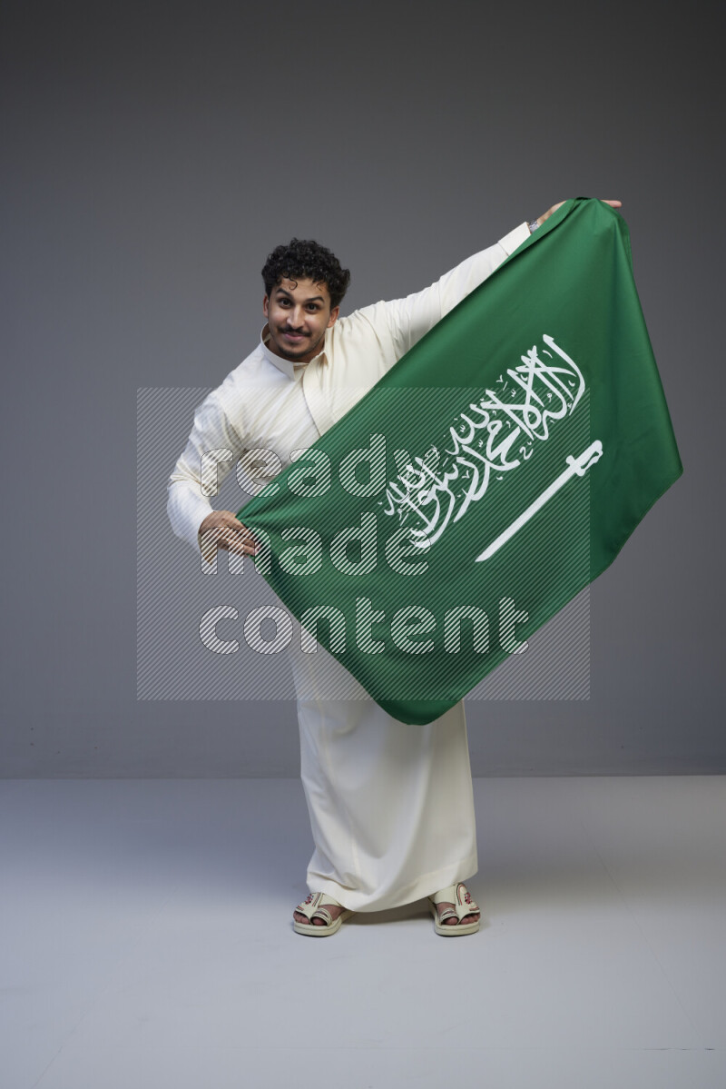 A Saudi man standing wearing thob holding big Saudi flag on gray background