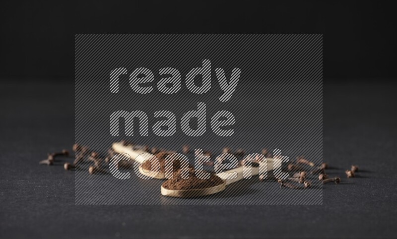 ملعقتان خشبيتان ممتلئتان ببودرة القرنفل مع حبوب القرنفل المتناثرة على خلفية سوداء