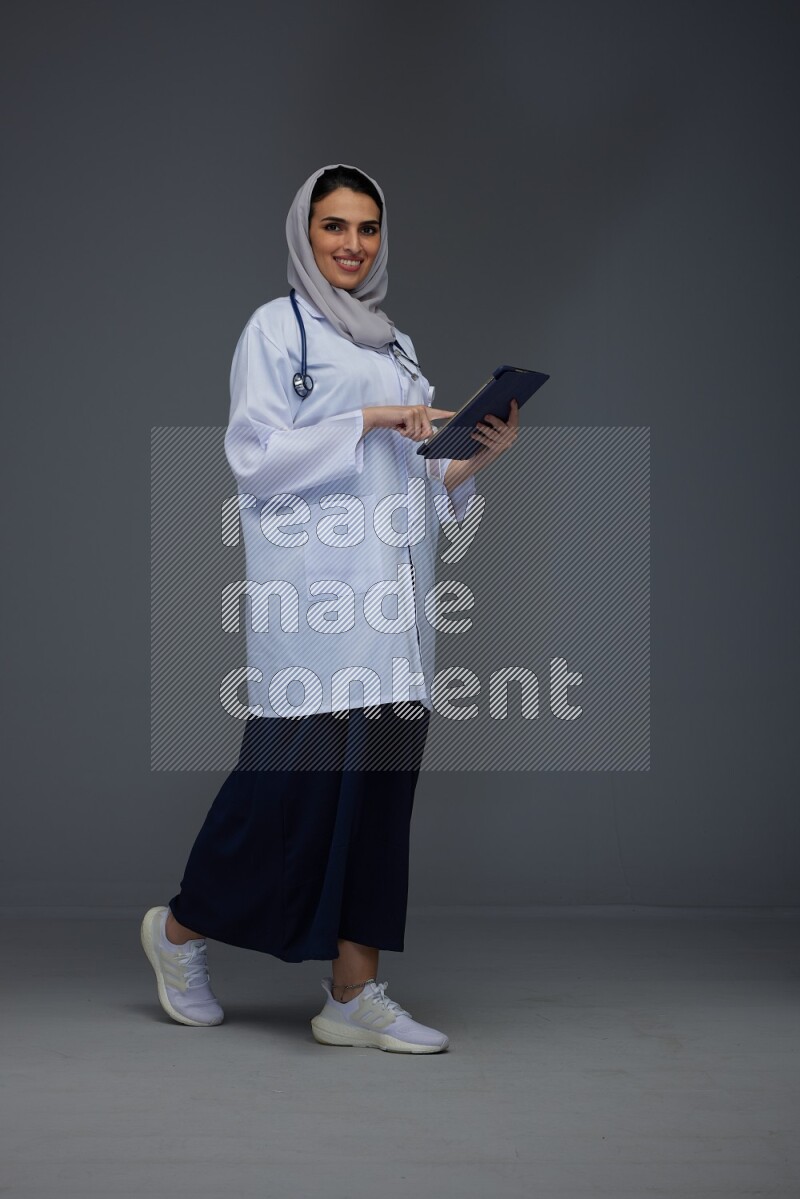 A Saudi doctor wearing a light gray head scarf standing and crossing her hands eye level on a grey background