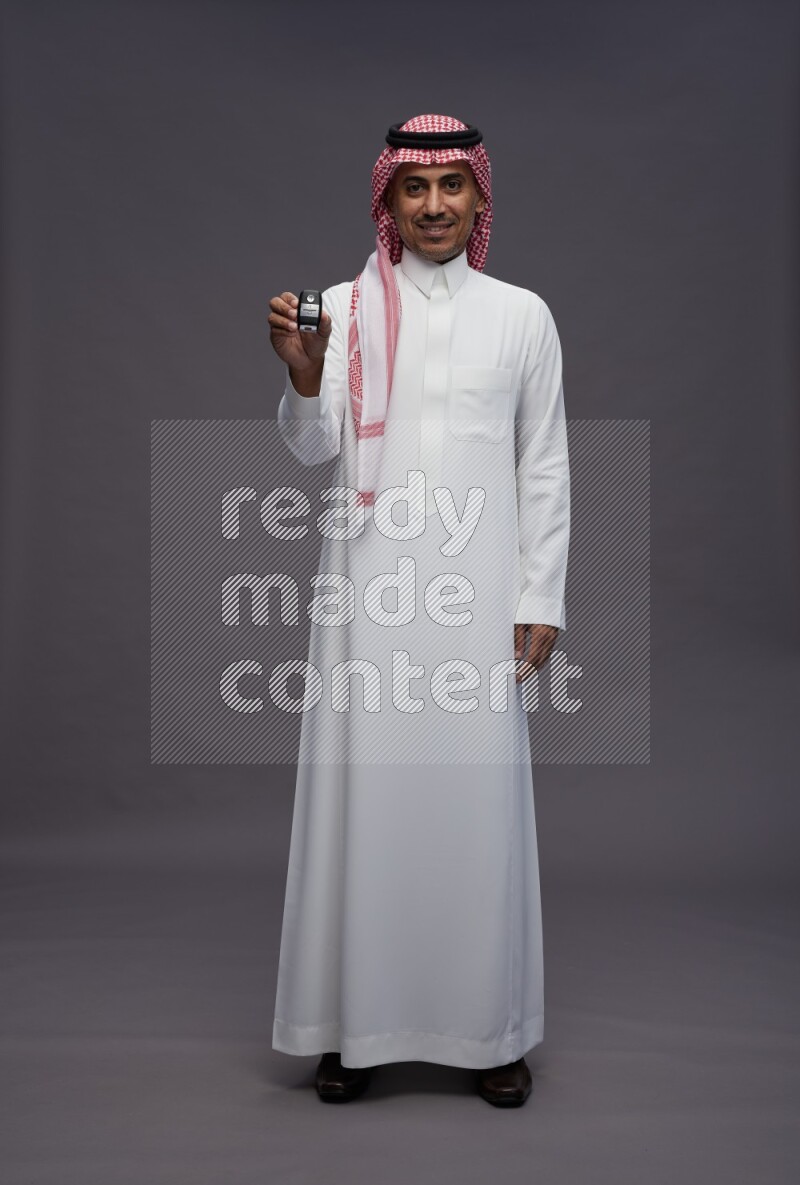 Saudi man wearing thob and shomag standing holding key car on gray background