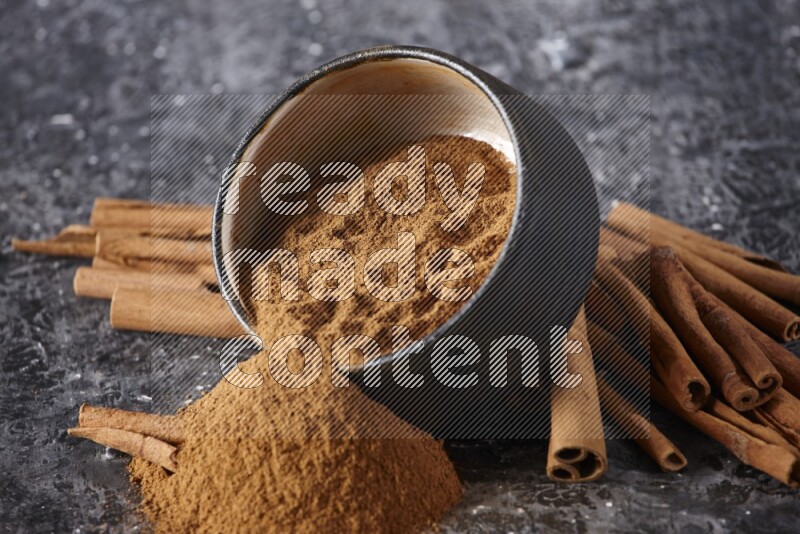 Black pottery bowl over filled with cinnamon powder and cinnamon sticks around the bowl on a textured black background