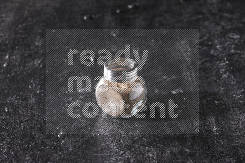 A glass spice jar full of black pepper powder on textured black flooring