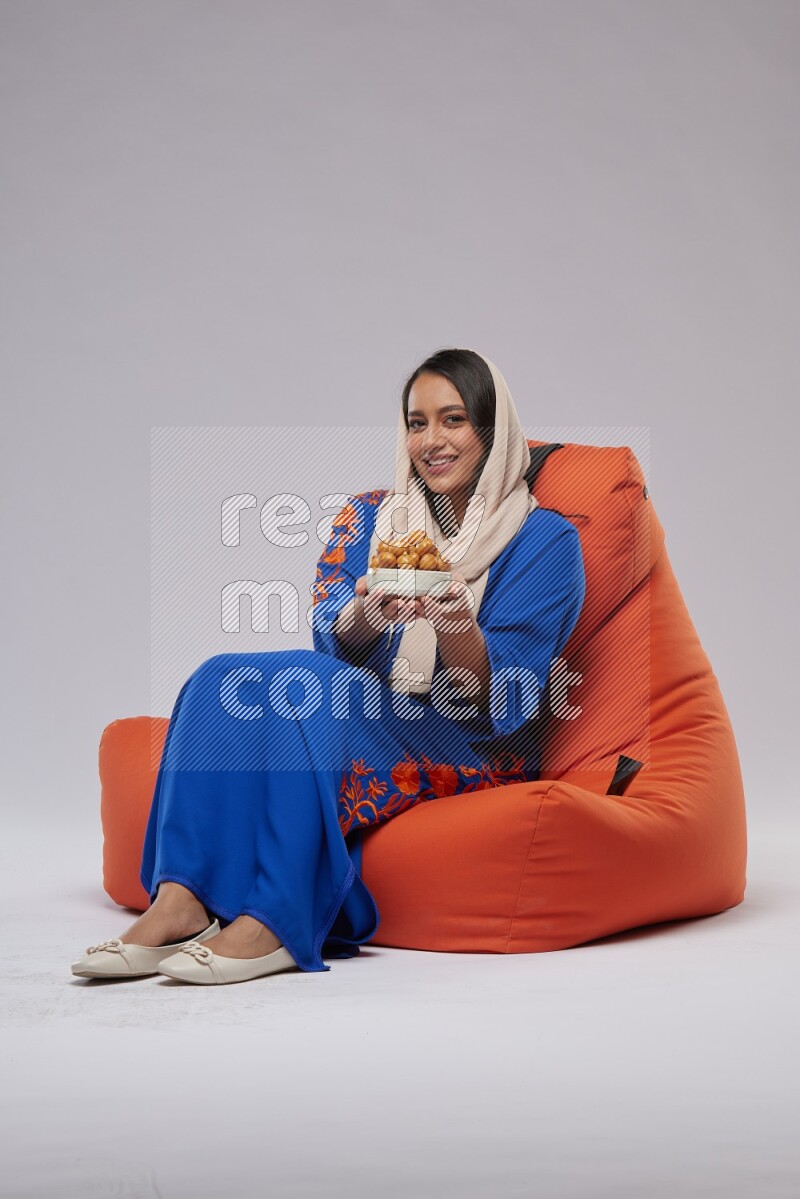 A Woman sitting on an orange beanbag wearing Jalabeya holding a plate of luqaimat