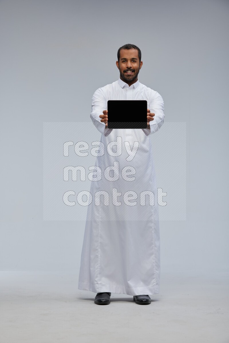 Saudi man wearing Thob standing showing tablet to camera on Gray background