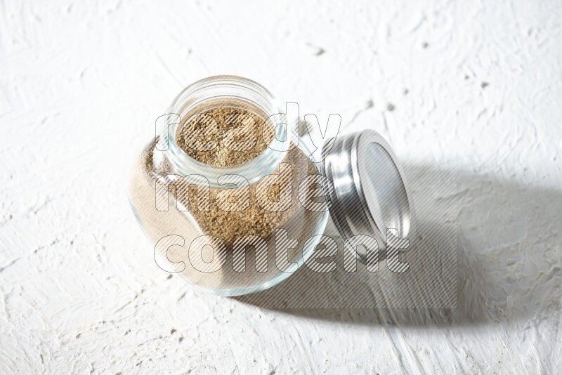 A glass spice jar full of cumin powder on textured white flooring