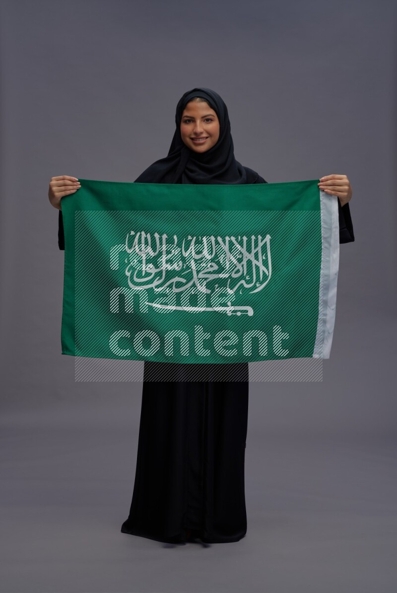 Saudi woman wearing Abaya standing holding Saudi flag on gray background