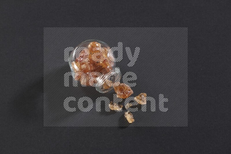 وعاء زجاجي ممتلئ بقطع الصمغ العربي والوعاء مقلوب وبعض القطع متناثرة خارجه علي خلفية سوداء