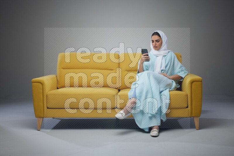 A Saudi woman wearing a light blue Abaya and white head scarf sitting on a yellow sofa and using her phone eye level on a grey background