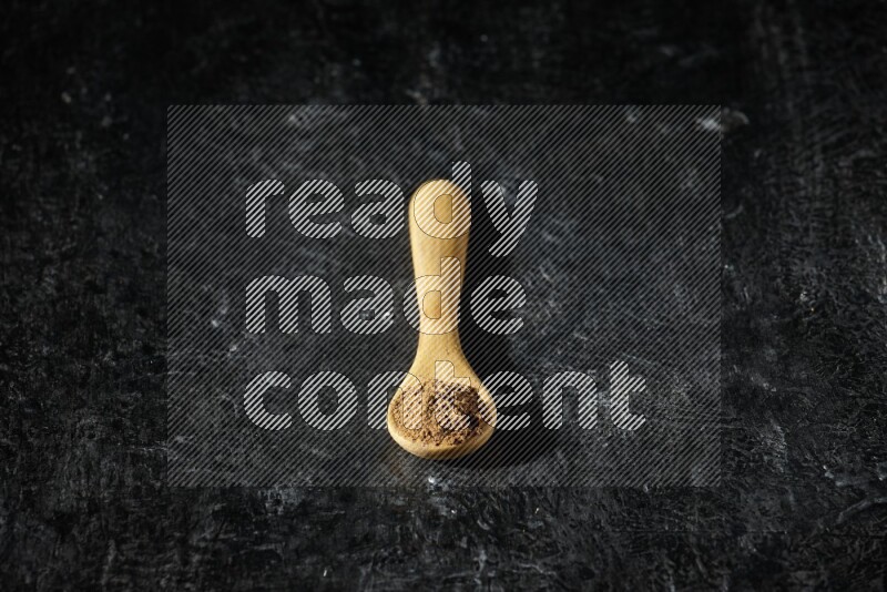 A wooden spoon full of allspice powder on a textured black flooring