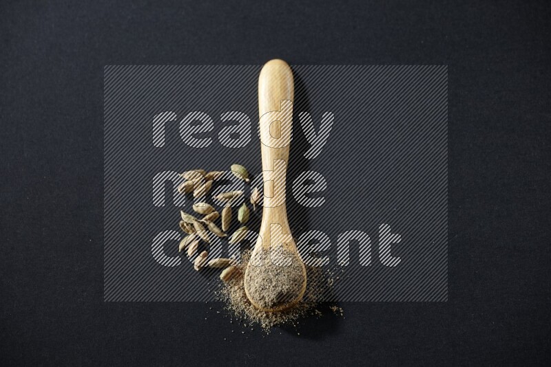 A wooden spoon full of cardamom powder and cardamom seeds beside it on black flooring