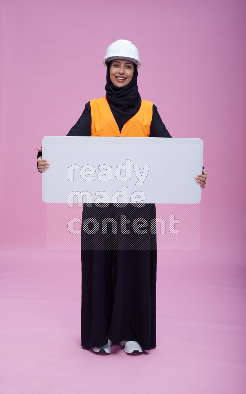 Saudi woman wearing Abaya with engineer vest and helmet standing holding board on pink background
