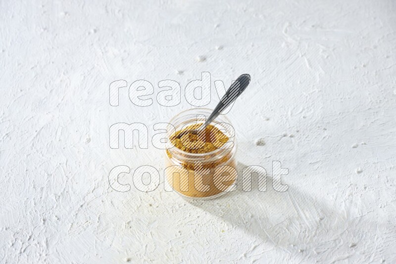 A glass jar and a metal spoon full of turmeric powder on a textured white flooring