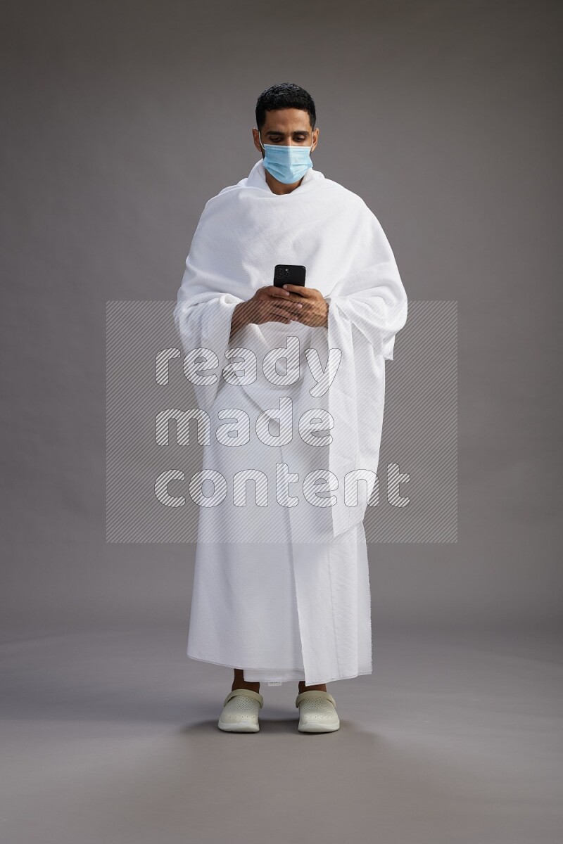A man wearing Ehram with face mask Standing texting on phone on gray background