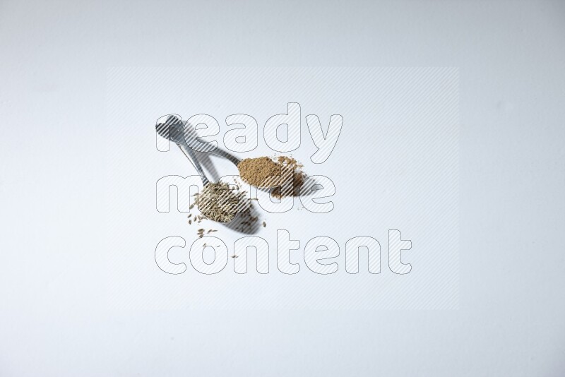 2 metal spoons full of cumin seeds and powder on white flooring