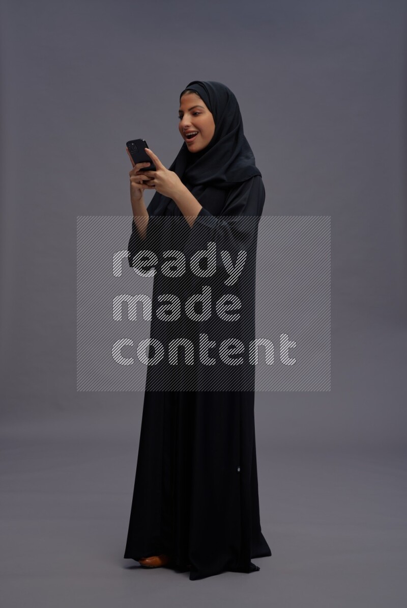 Saudi woman wearing Abaya standing texting on phone on gray background