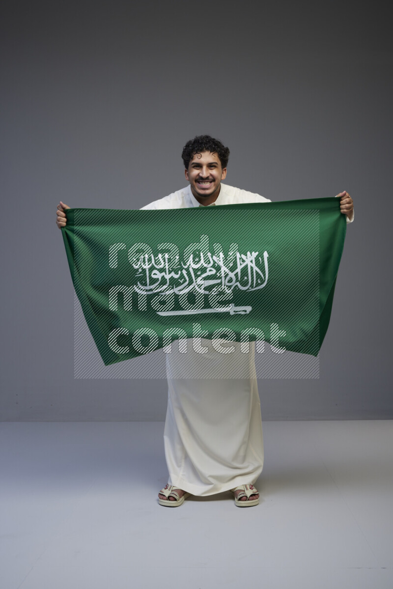 A Saudi man standing wearing thob holding big Saudi flag on gray background