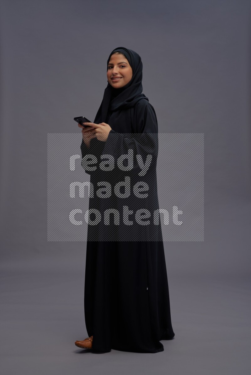 Saudi woman wearing Abaya standing texting on phone on gray background