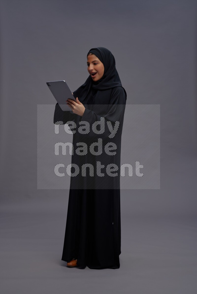 Saudi woman wearing Abaya standing working on tablet on gray background