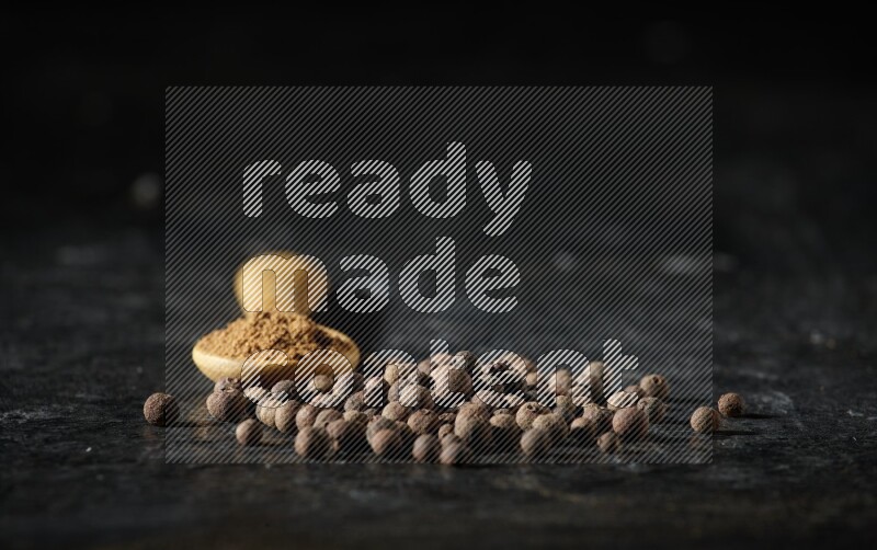 A wooden spoon full of allspice powder and whole balls spreaded on a textured black flooring