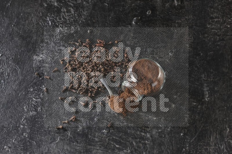 A flipped glass spice jar and a metal spoon full of cloves powder with cloves spread on textured black flooring