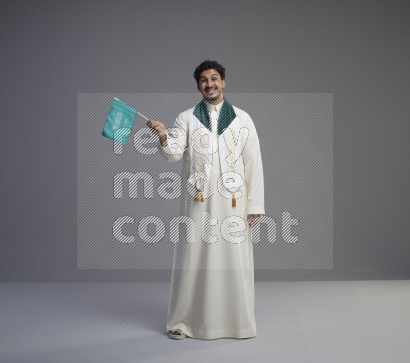 A Saudi man standing wearing thob and saudi flag scarf and holding small saudi flag on gray background