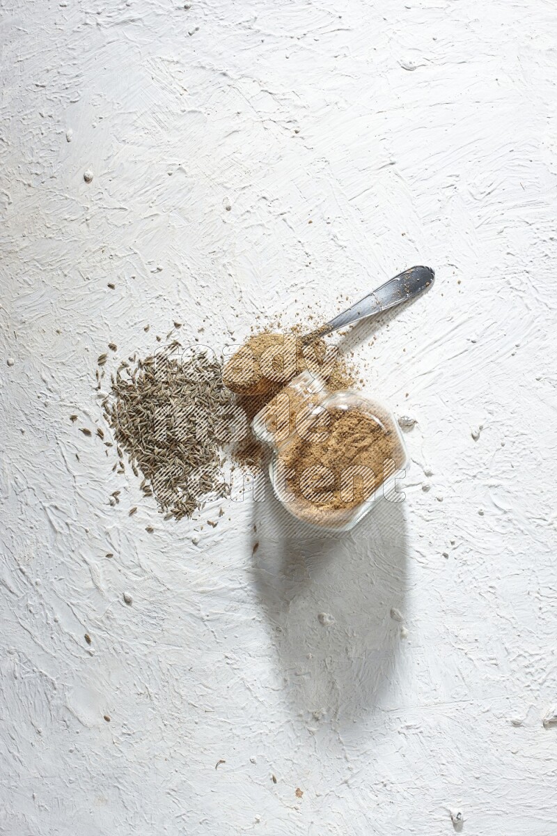 A glass spice jar and metal spoon full of cumin powder and the jar flipped and powder spilled out with cumin seeds on textured white flooring