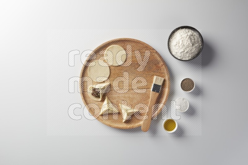 two closed sambosas and one open sambosa filled with meat while flour, salt, black pepper and oil with oil brush aside in a wooden dish on a white background