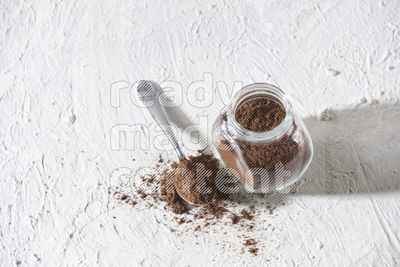 A glass spice jar and a metal spoon full of cloves powder on textured white flooring