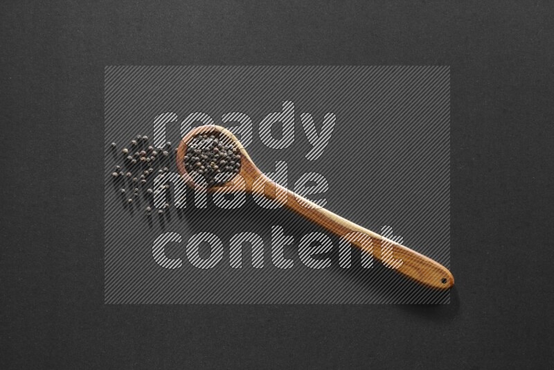A wooden ladle full of black pepper beads on a black flooring