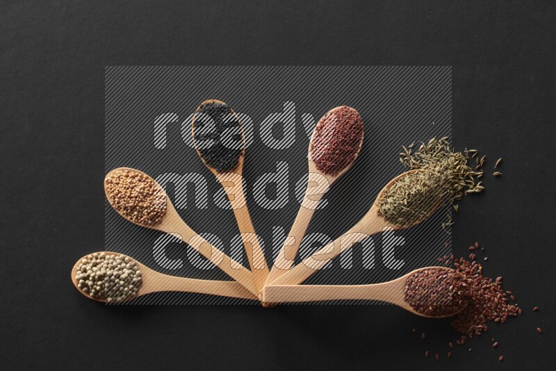 wooden spoons filled with white peppers, mustard seeds, black seeds, garden cress, cumin and flax on black flooring