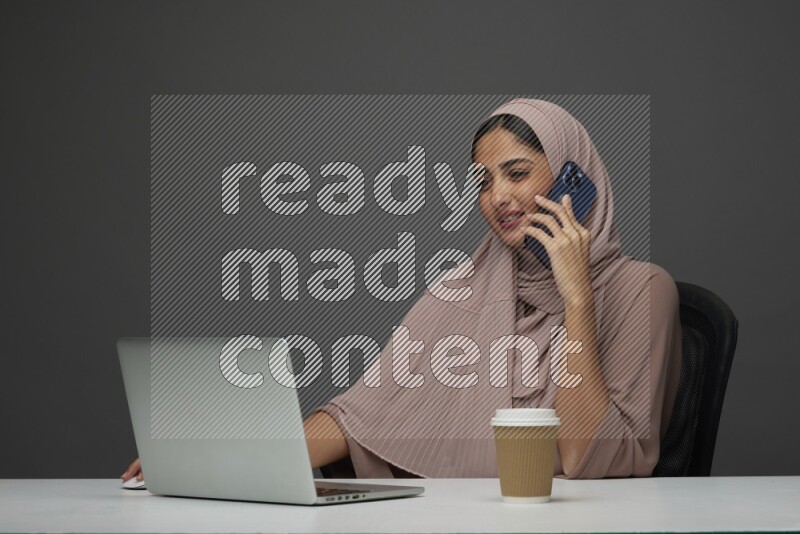A Saudi woman Setting on her desk
 calling  on a Gray Background wearing Brown Abaya with Hijab