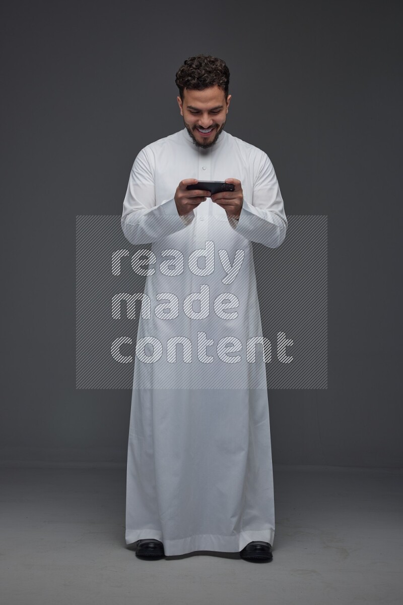 A Saudi man wearing Thobe and playing using his phone horizontally playing video games while standing and making different poses eye level on a gray background