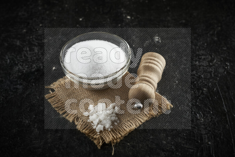 وعاء زجاجي ممتلئ بالملح الأبيض مع مطحنة خشبية على قماش الخيش على خلفية سوداء