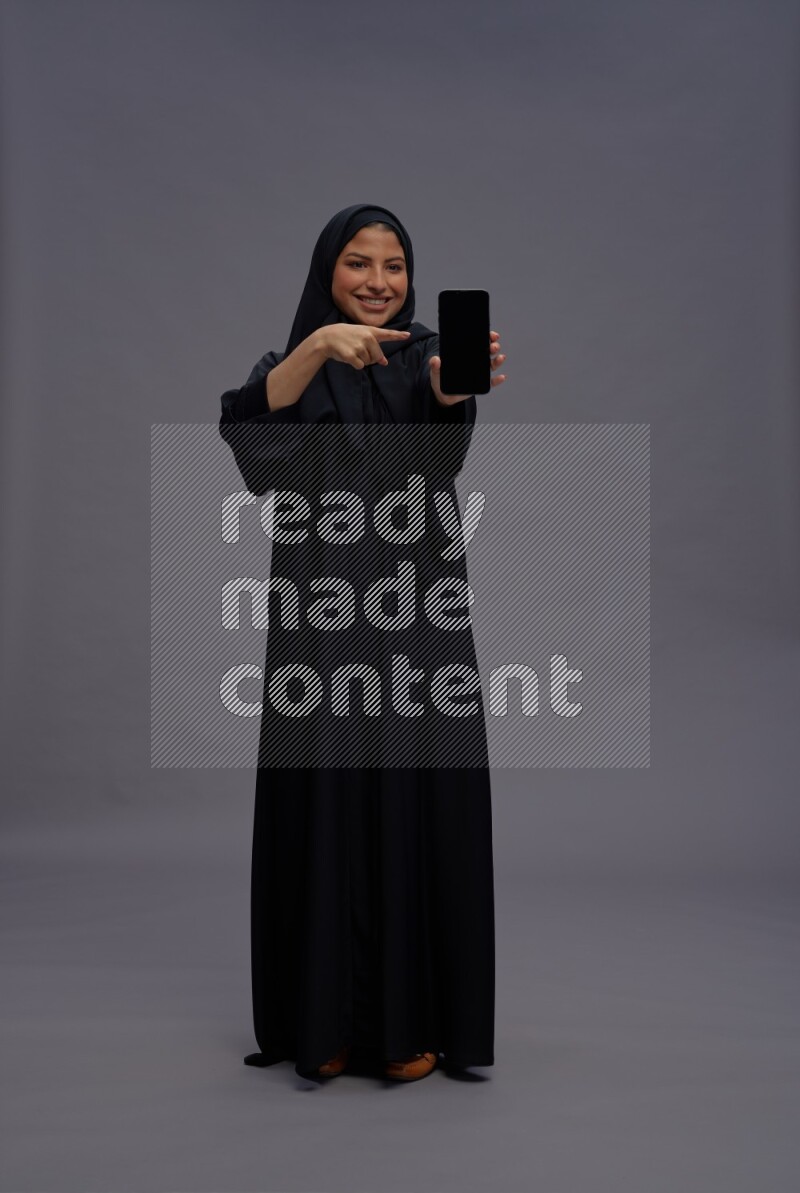 Saudi woman wearing Abaya standing showing phone to camera on gray background