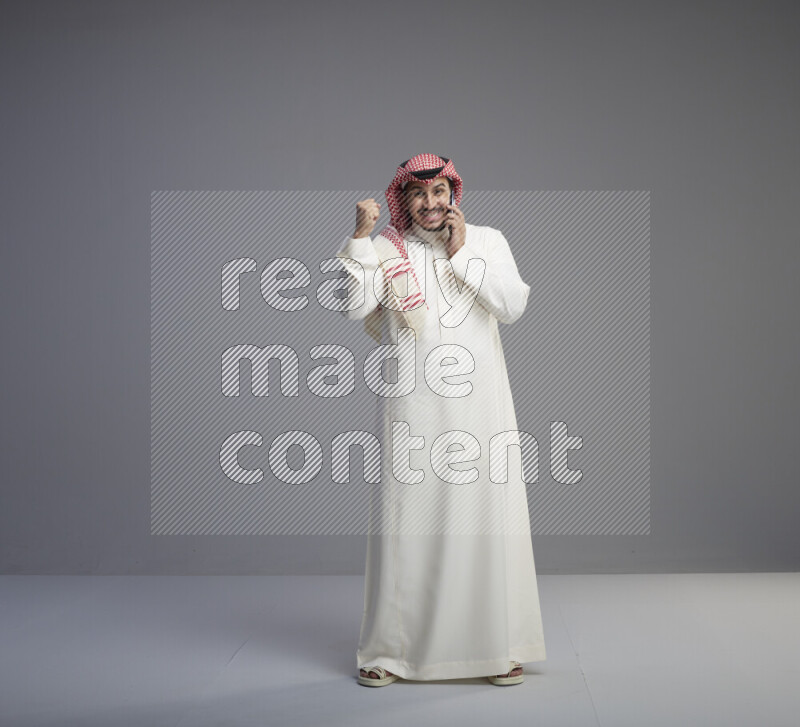 A Saudi man standing wearing thob and red shomag talking on phone on gray background