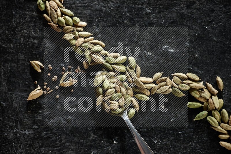 A Metal spoon full of cardamom seeds and some seeds beside it on a textured black flooring